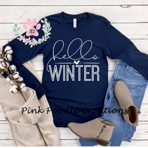 May include: Navy blue long-sleeve shirt with the words "hello WINTER" in white script. The shirt is paired with blue jeans, tan booties, and a beige cardigan. A sprig of cotton and a floral design are also in the image.