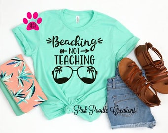 May include: A turquoise t-shirt with the text "Beaching - Not - Teaching" and a pair of sunglasses with palm trees on the lenses. The shirt is laid out on a table with a pair of brown sandals and denim shorts.