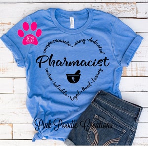 May include: A blue t-shirt with a black heart design that says "Pharmacist" with a mortar and pestle inside the heart. The words "compassionate, caring, dedicated, warm, reliable, loyal, kind, loving" surround the heart. A pink paw print with the letters "SG" inside is in the upper left corner. The t-shirt is on a white wooden surface.