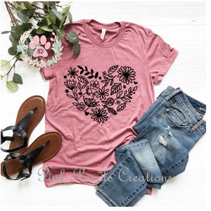 May include: A pink t-shirt with a black floral heart design. The shirt is laid out on a white surface with a pair of brown sandals and a pair of blue jeans.