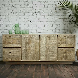 May include: A light-coloured wooden cabinet with carved geometric patterns on the doors and drawers. The cabinet has a green jug and glasses on top. A large potted plant is to the right.