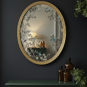 May include: An oval mirror with a gold frame featuring a decorative painted design of branches, leaves, small blue and white flowers, and birds. The floral and avian motif is applied directly onto the glass surface, creating a detailed border.