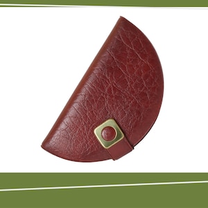 May include: A burgundy leather case with a semi-circular shape. The case has a textured surface and a gold-colored metal clasp. The case is designed to hold small items and is set against a white background.