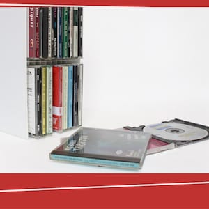 May include: A selection of CDs in their cases, some stored in a white unit, others lying open. The CD cases show different album titles and colours, highlighting music media.