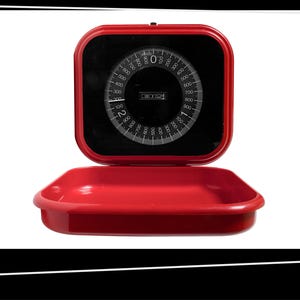 May include: A vintage red darkroom timer with a black face and white markings. The timer has a circular dial with numbers and a rectangular base. The timer is open, revealing the dial and the base.
