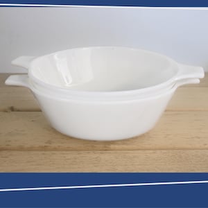 May include: Two white ceramic bowls with handles, stacked on top of each other. The bowls are on a light brown wooden surface.