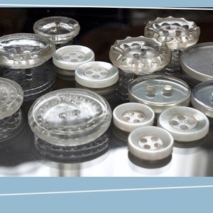 May include: A collection of vintage buttons in various shapes, sizes, and colors, including clear glass, white plastic, and silver metal. Some buttons have a raised design, while others are smooth.