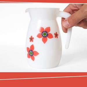 Op de afbeelding: Witte kan met een handvat en een bloemenmotief. De bloemen zijn rood met blauwe en groene centra.