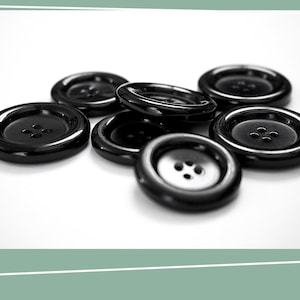 May include: A group of black plastic buttons with four holes each. The buttons are arranged in a pile on a white surface.
