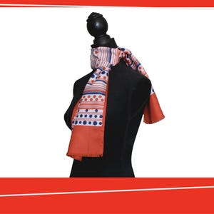 Pode incluir: Um len&ccedil;o estampado com um design vermelho, branco e azul est&aacute; drapeado sobre um manequim preto. O len&ccedil;o apresenta listras, pontos e uma borda com franjas. &Eacute; um acess&oacute;rio de moda.