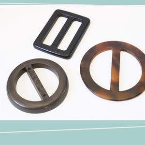 May include: Three belt buckles in different shapes and colors. A black rectangular buckle with two slots, a dark brown circular buckle with a single bar, and a tortoiseshell-patterned circular buckle with a single bar.