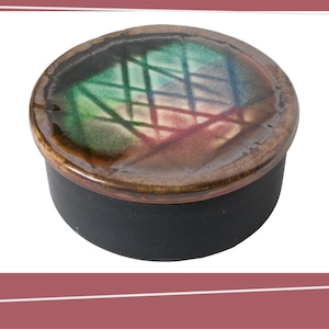 Op de afbeelding: Een ronde keramische sieradendoos met een deksel. Het deksel heeft een kleurrijk abstract ontwerp in groen, rood en blauw, met een rasterpatroon. De doos zelf is zwart met een bruine rand. De doos wordt waarschijnlijk gebruikt voor het opbergen van kleine items.