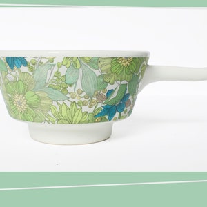 Op de afbeelding: Een witte keramische kom met een bloemenpatroon in tinten groen, blauw en geel. De kom heeft een handvat en is perfect om soep of salade te serveren.