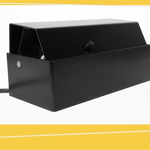 May include: A black metal desk lamp with a rectangular base and a sloped top. The lamp has a small knob on the side and a power cord. The lamp is designed to direct light downwards.