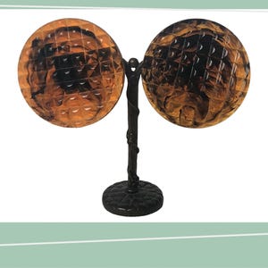 Op de afbeelding: Een decoratieve tafellamp met twee cirkelvormige, amberkleurige kappen, elk met een textuurpatroon. De kappen zijn verbonden door een donkere, takachtige steel en een ronde voet. De lamp is ontworpen voor sfeerverlichting.