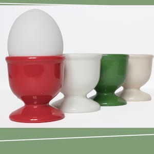 Op de afbeelding: Een wit ei in een rode keramische eierdop, met drie andere eierdoppen in wit, groen en crème. De eierdoppen staan in een rij, wat hun eenvoudige ontwerp benadrukt. De achtergrond is wit.