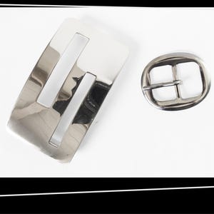 May include: Two silver-toned metal belt buckles. One is rectangular with two parallel cutouts, the other is oval with a single bar across the center. Both have a shiny, reflective surface.