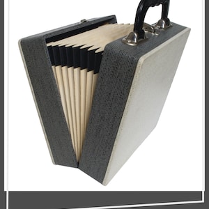 May include: A portable expanding file folder with a textured dark gray exterior and a light cream-colored side panel. The case features a black plastic handle with silver-toned metal hardware and contains multiple interior dividers in a light tan color.