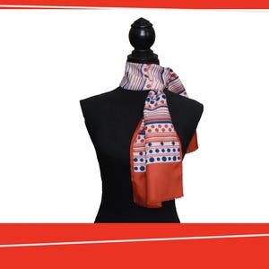 May include: A silk scarf with a geometric pattern of orange, white, and blue stripes and dots. The scarf is draped over a black mannequin, showcasing its design and color scheme. The scarf's edges are finished with a simple hem.