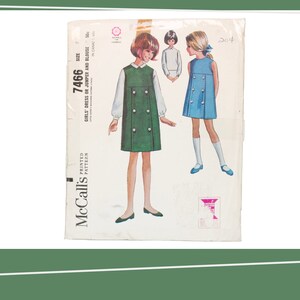 Op de afbeelding: Vintage McCall's naaipatroon voor een meisjesjurk of trui en blouse. Het patroonnummer is 7466 en de maat is 7. Het patroon toont een groene jurk met witte knopen en een blauwe jurk met witte knopen.