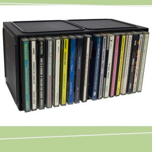 May include: A black CD storage case containing numerous CDs. The CDs display diverse album artwork and text, with titles such as "Power of Lard EP" and "Follow the Leader". The case features a hinged lid.