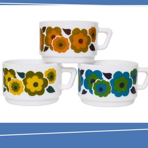 Puede incluir: Tres tazas de cerámica blanca con diseños florales en naranja, amarillo y azul. Cada taza tiene un asa pequeña y un diseño floral repetido alrededor del borde. Las tazas están apiladas, mostrando los diferentes esquemas de color.