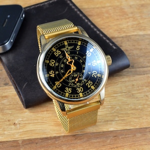 May include: A gold-tone watch with a black face and a mesh metal band. The watch face has white markings and a small second hand. The watch is on a wooden surface.