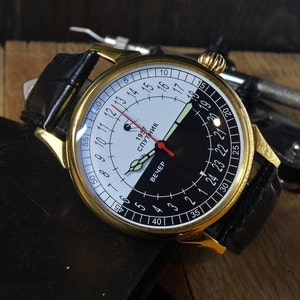 May include: A gold-tone wristwatch with a black leather band. The watch face is white and black with a red hand and the text "1957 СПУТНИК" and "ВЕЧЕР".