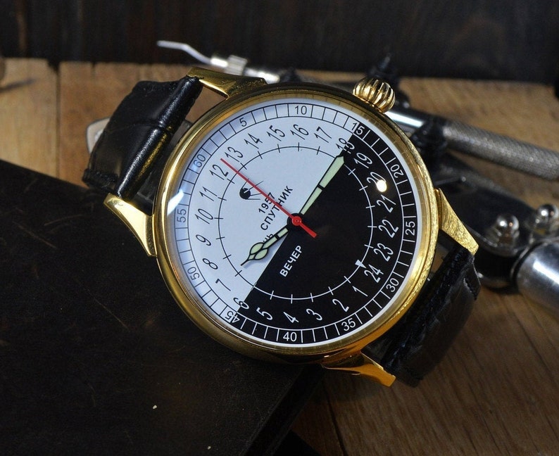 May include: A gold-tone wristwatch with a black leather band. The watch face is white and black with a red hand and the text "1957 СПУТНИК" and "ВЕЧЕР".