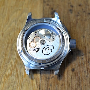May include: The back of a silver wristwatch with a clear caseback revealing the inner workings of the watch. The watch has the text "+ AMOVIE" and "PRO1780D" printed on the caseback.