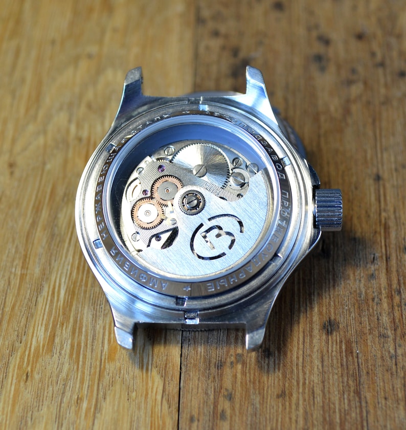 May include: The back of a silver wristwatch with a clear caseback revealing the inner workings of the watch. The watch has the text "+ AMOVIE" and "PRO1780D" printed on the caseback.