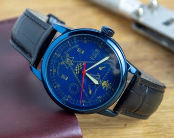 Puede incluir: Un reloj azul con una correa de cuero negro. La esfera del reloj tiene marcas doradas y una aguja de segundos roja. La esfera del reloj también tiene un símbolo masónico.