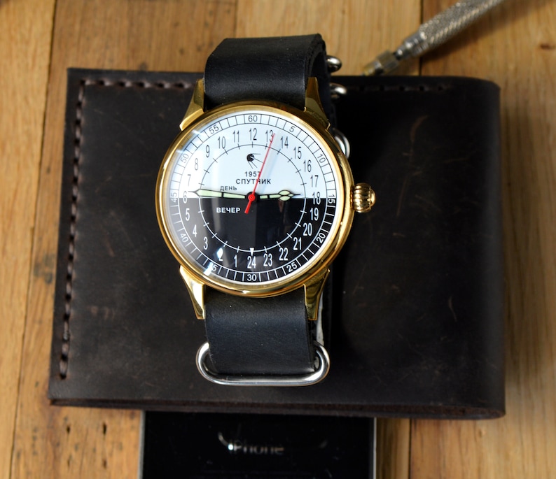 May include: A gold-tone wristwatch with a black leather band. The watch face is white with black markings and features the text "1957 Спутник" and "День Вечер".
