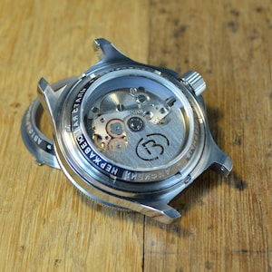 May include: A silver watch with a see-through back revealing the inner workings of the watch. The watch face has the text "ARCTIC" and "НЕПРАВДА" in Cyrillic script.