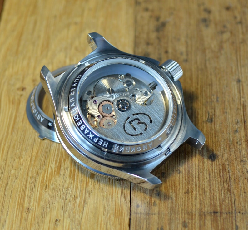 May include: A silver watch with a see-through back revealing the inner workings of the watch. The watch face has the text "ARCTIC" and "НЕПРАВДА" in Cyrillic script.