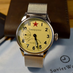 Può includere: Un orologio da polso argentato con un quadrante giallo e numeri neri. L'orologio ha una stella rossa e il testo "Командирские" sul quadrante. L'orologio è su un cinturino in maglia argentata.