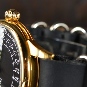 May include: A gold-tone wristwatch with a black and white face. The face has a 24-hour clock with the numbers 9 through 18 on the white side and 19 through 24 on the black side. The watch has a black leather strap with silver buckles.