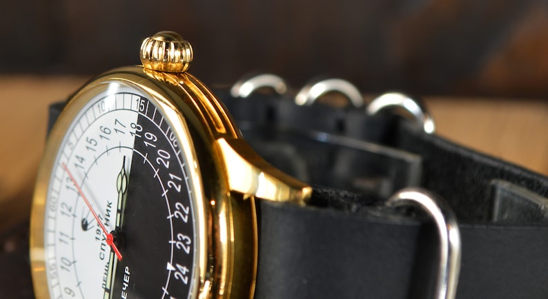 May include: A gold-tone wristwatch with a black and white face. The face has a 24-hour clock with the numbers 9 through 18 on the white side and 19 through 24 on the black side. The watch has a black leather strap with silver buckles.