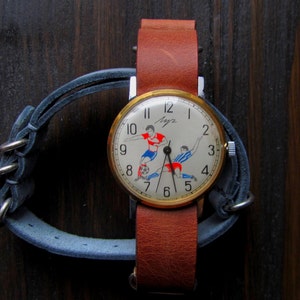 May include: A vintage wristwatch with a white face and black hands. The watch face features a red, blue, and white illustration of two soccer players. The watch has a brown leather band.