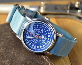 Puede incluir: Un reloj de pulsera con caja redonda plateada y esfera azul con un patrón de cuadrícula blanco, marcadores de 24 horas y texto en cirílico. El reloj tiene una correa de cuero azul claro, un segundero rojo y manecillas blancas.