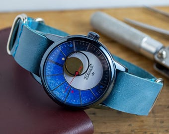 May include: A wristwatch with a blue leather strap and a black case. The watch face has a distinctive design with blue and white segments, a gold circle, and a red second hand. The watch is labelled "Takema".