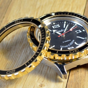 May include: A gold-tone watch with a black face and a black bezel. The watch has a date window at the 3 o'clock position and the text "31 КАМЕНЬ СДЕЛАНО В РОССИЯ" on the face. The watch is shown next to a spare black bezel.