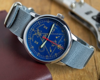 Puede incluir: Un reloj de pulsera plateado con una esfera azul y detalles dorados. La esfera presenta un diseño de brújula con una aguja de segundos roja. El reloj tiene una correa de cuero gris.