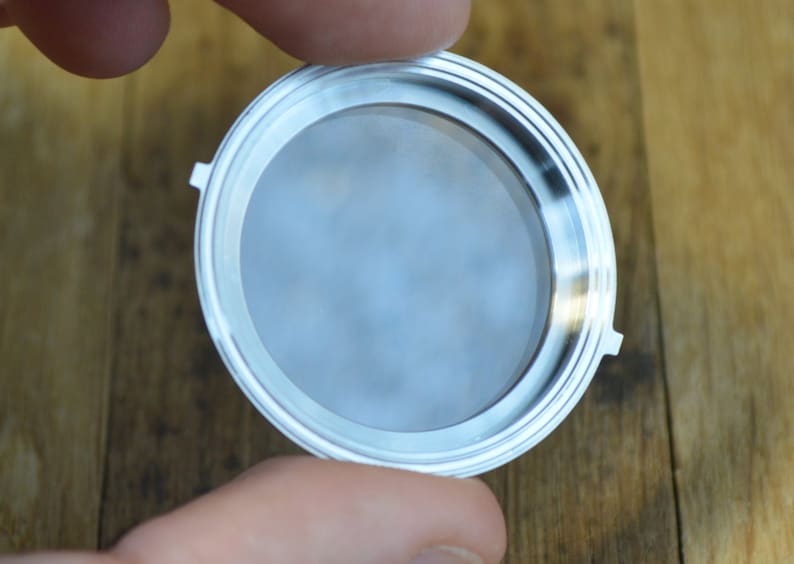 May include: A silver-colored metal filter with a round, flat base and a raised rim. The filter is designed to fit into a coffee maker.