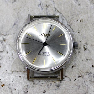 May include: A silver wristwatch with a white face and black hands. The watch face has gold accents and the text "23 jewels Shockproof".