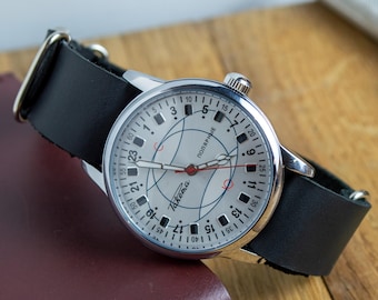 May include: A silver wristwatch with a white face, black numerals, and red detailing. The watch has a black leather strap. The watch face displays the word "Takema" and the Cyrillic word "ПОЛЯРНЫЕ".