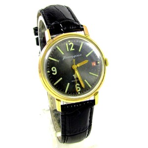 Peut inclure: Une montre-bracelet dorée avec un cadran noir et un bracelet en cuir noir. Le cadran de la montre a des aiguilles et des marqueurs dorés, et le texte "Радиочасы" est imprimé en blanc.
