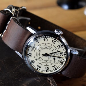 May include: A silver wristwatch with a white face and black markings. The watch has a brown leather strap with a buckle closure. The watch face has a small airplane symbol and the numbers 5, 10, 15, 20, 25, 30, 35, 40, 45, 50, 55, and 60.