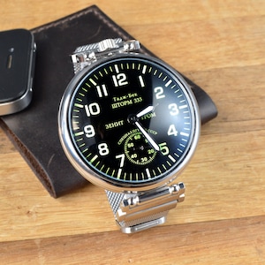 Può includere: Un orologio da polso argentato con quadrante nero e cinturino in metallo a rete. Il quadrante dell'orologio ha numeri e lancette bianchi, e il testo "Тадж-Бек ШТОРМ 333" e "ЗЕНИТ ТРОМ СССР" in caratteri cirillici.