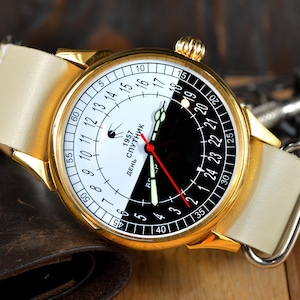 May include: A gold-tone wristwatch with a white and black face. The face has a 24-hour clock with the text "День Спутник 1957" (Day Satellite 1957). The watch has a white leather band.